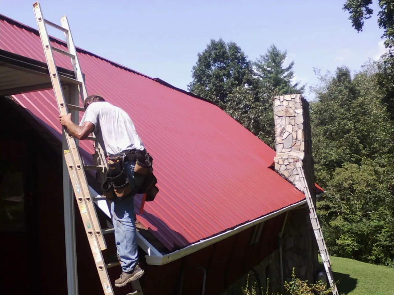 Roofer on a ladder beside a red metal roof and stone chimney — Soffit & Fascia Repair by Roofing Brothers in Metaline Falls